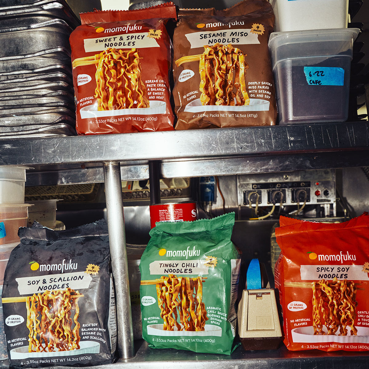 Multiple bags of Momofuku instant noodles arranged on a metal kitchen shelf, including Sweet & Spicy, Sesame Miso, Soy & Scallion, and Tingly Chilli Noodles.